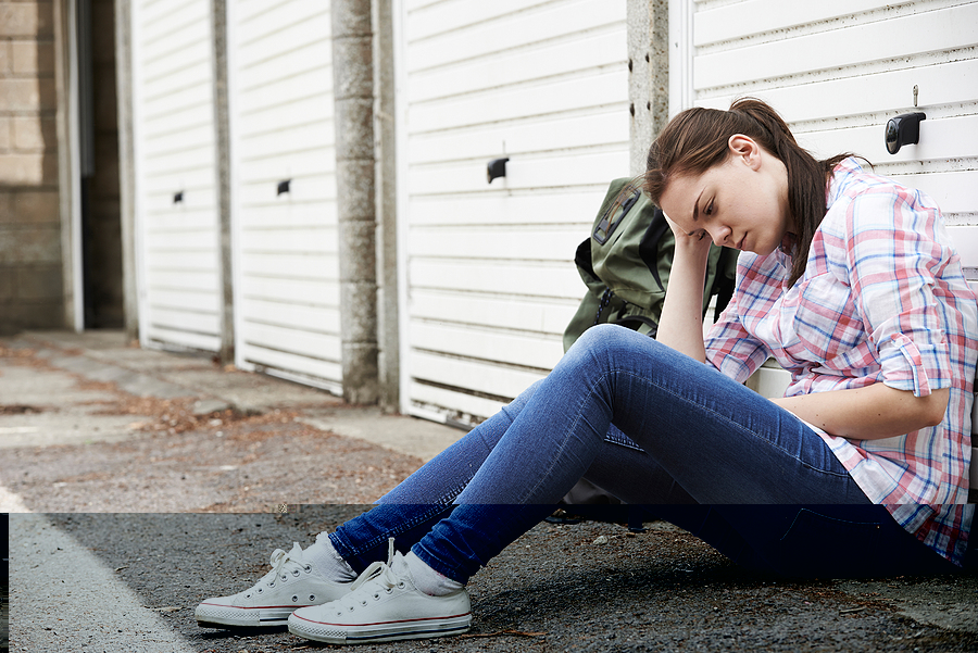 Homeless teen girl with backpack sitting out on the sidewalk.