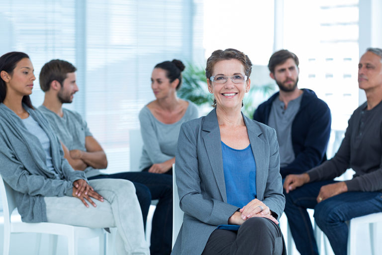 Portrait of a smiling female therapist with group therapy in session in background